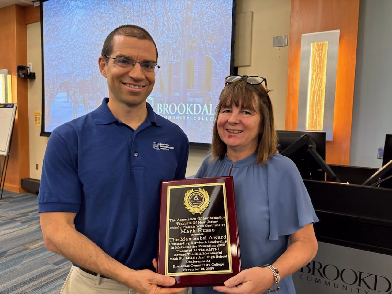 Two people stand indoors at Brookdale Community College. The man holds a plaque that reads “The Max Sobel Award” as both smile at the camera. Presentation slides and classroom equipment are visible in the background.